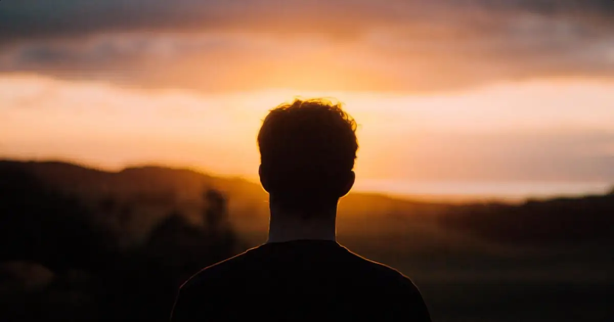 Foto de garoto de costas olhando para o sol se por no horizonte.